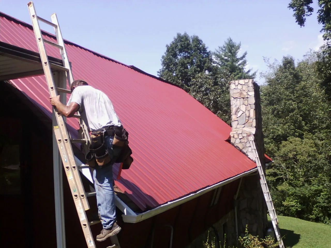 Roofer on a ladder beside a red metal roof and stone chimney — Soffit & Fascia Repair by Roofing Brothers in Shady Side